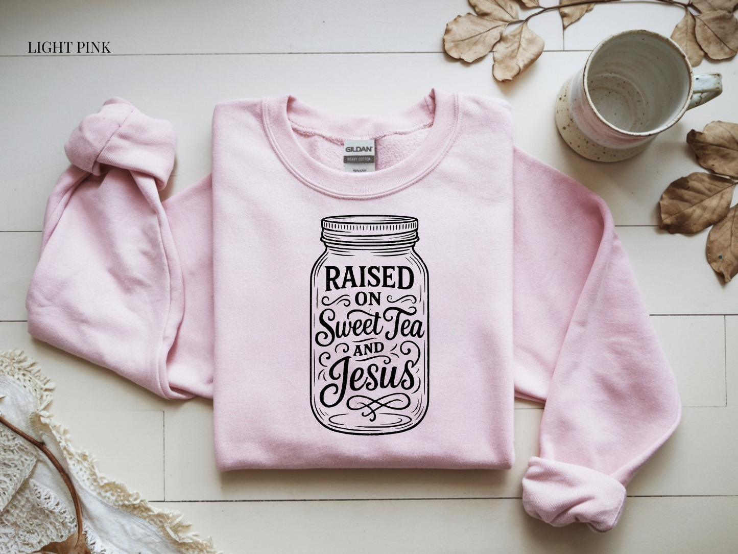 Pink sweatshirt with 'Raised on Sweet Tea and Jesus' design on a light wooden surface.