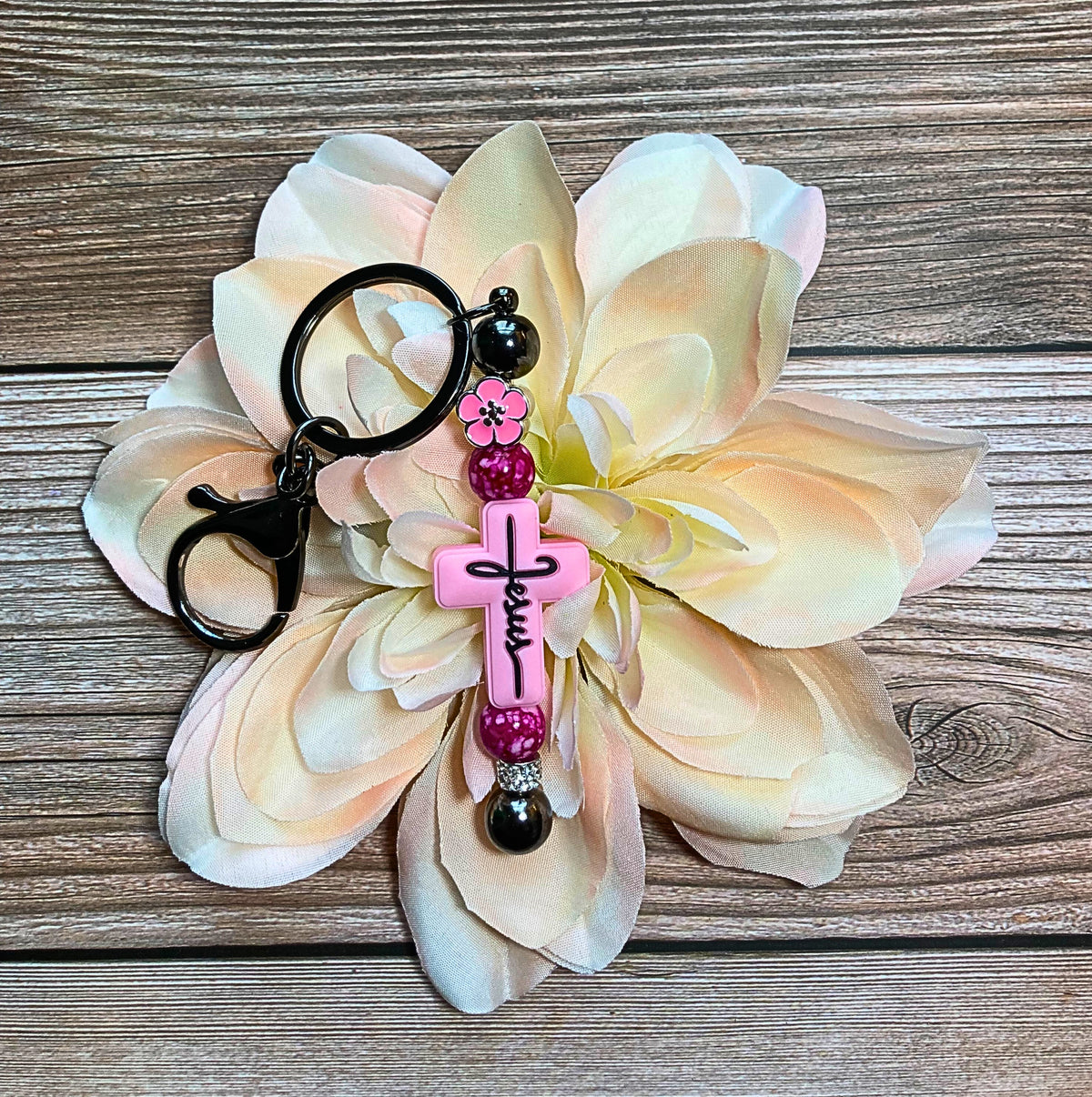 A beadable keychain with a pink cross focal bead and multicolored accent beads, displayed on a artificial flower against a wooden background.