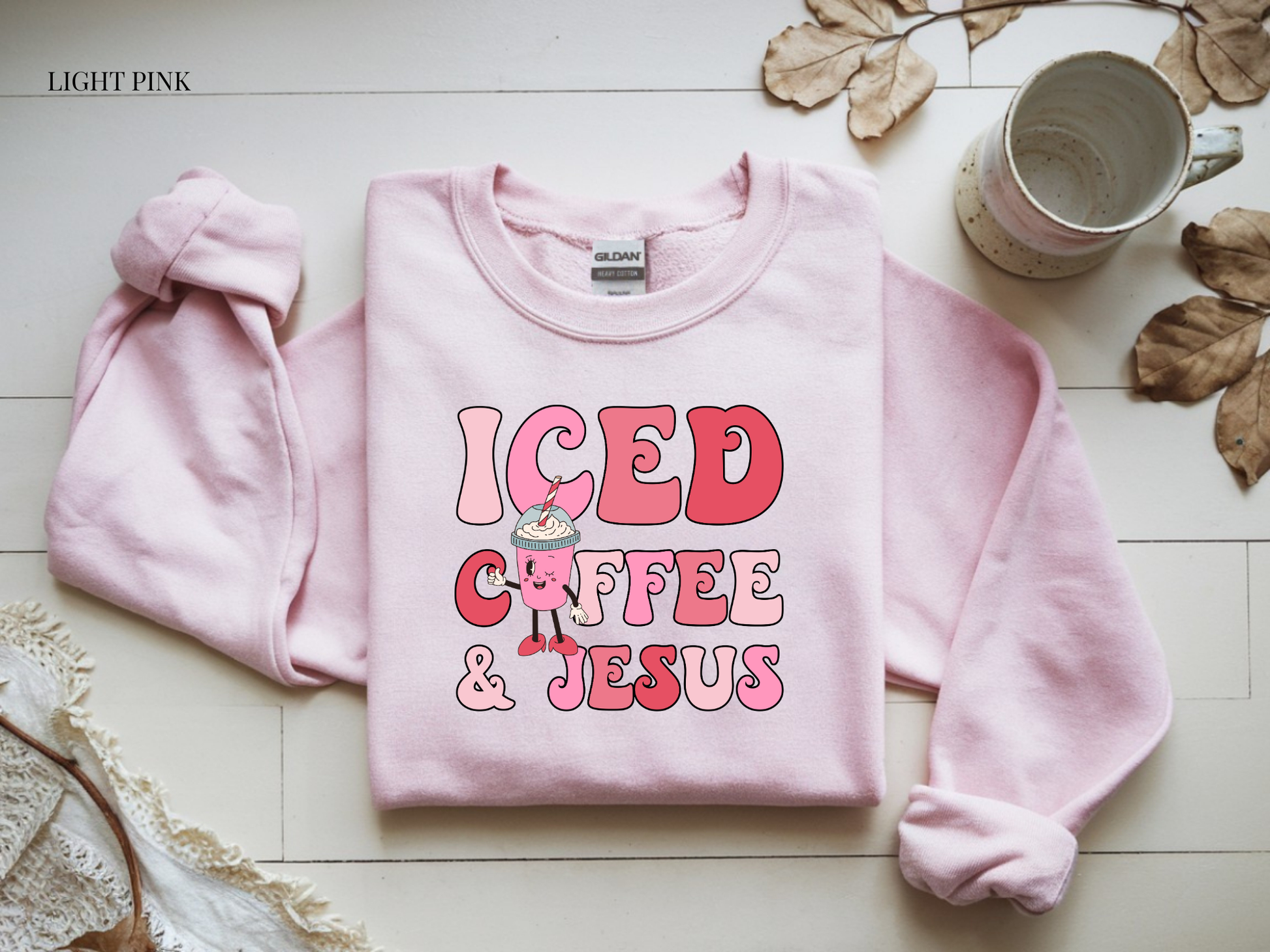 Light pink sweatshirt with 'ICED COFFEE & JESUS' text on a light wooden surface.