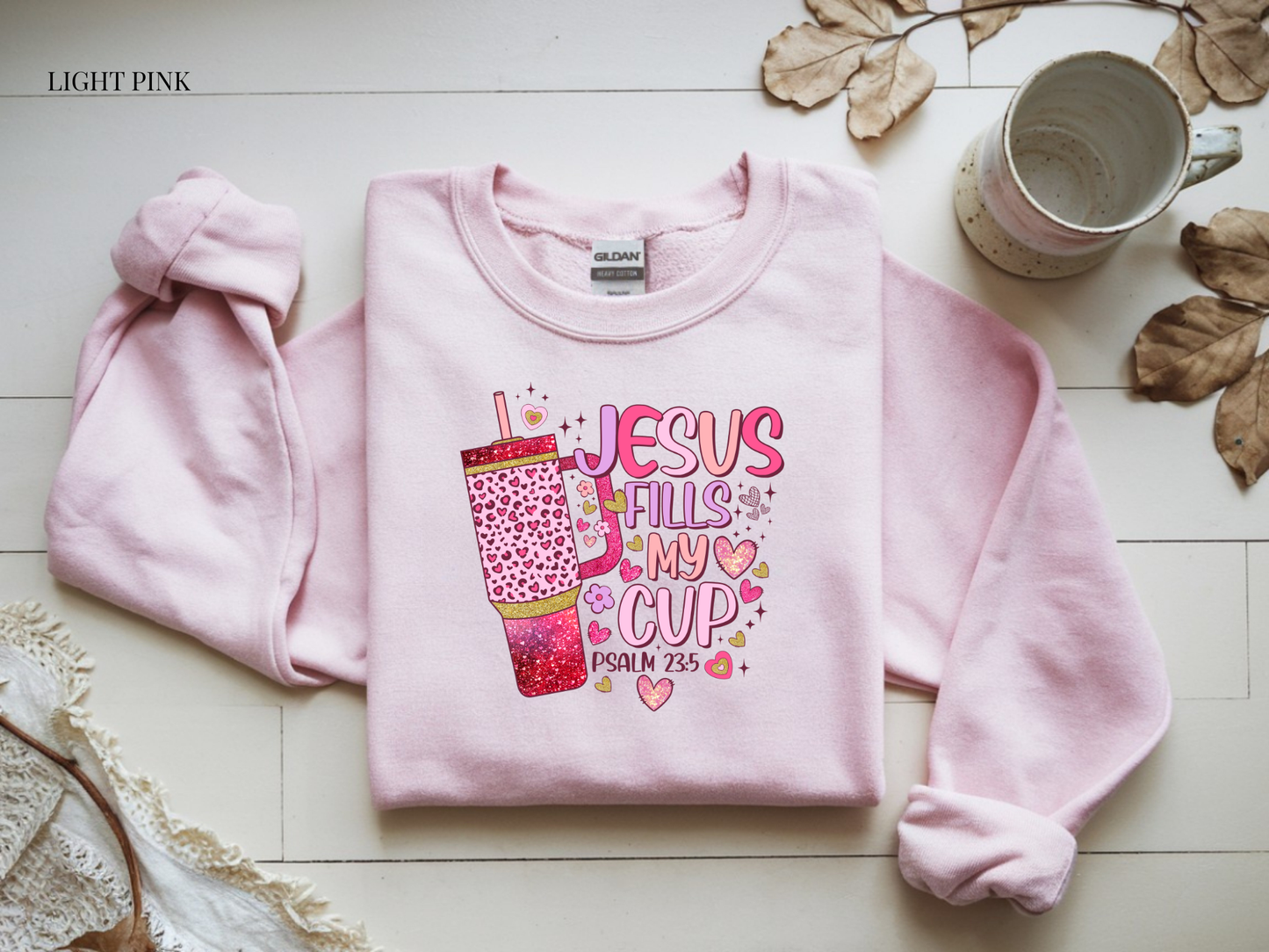 Light pink sweatshirt with 'Jesus Fills My Cup' design on a light wooden surface.