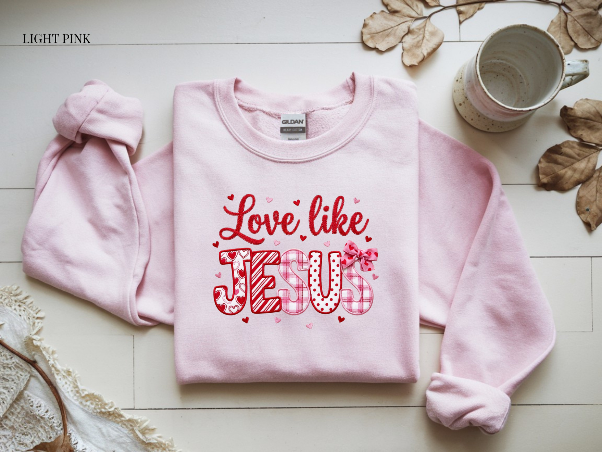 Light pink sweatshirt with 'Love like JEUX' text on a white surface with leaves and a mug.