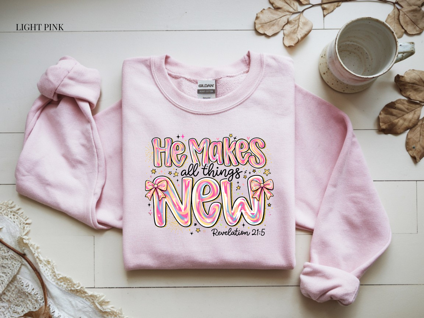 Light pink sweatshirt with colorful text on a wooden surface with leaves and a mug.