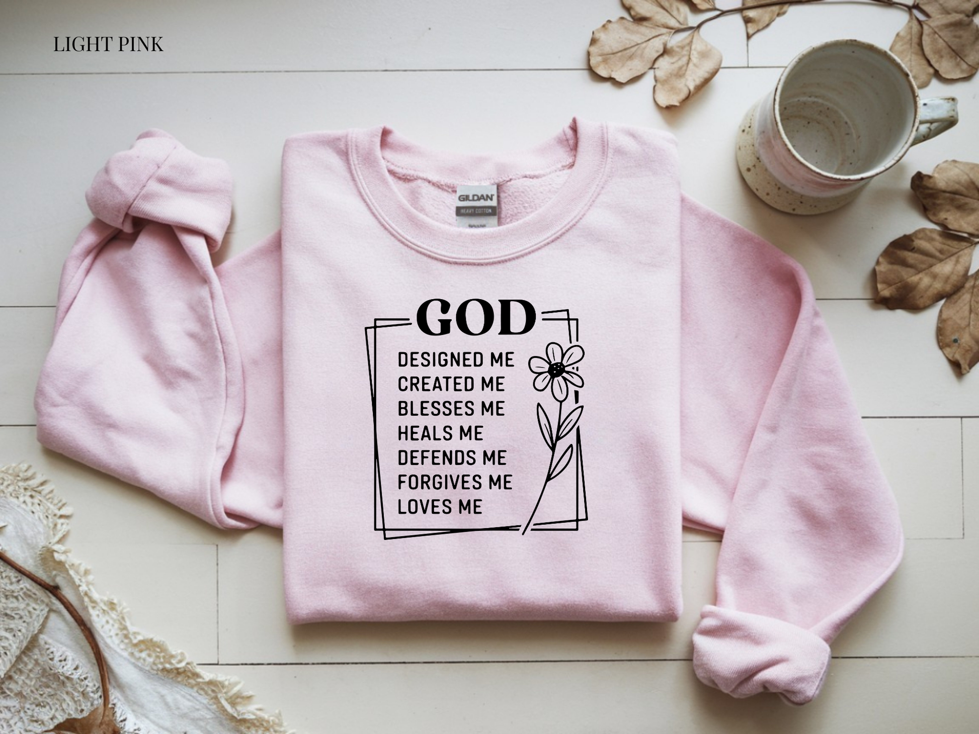 Pink sweatshirt with motivational quote on a light wooden surface with a mug and leaves.