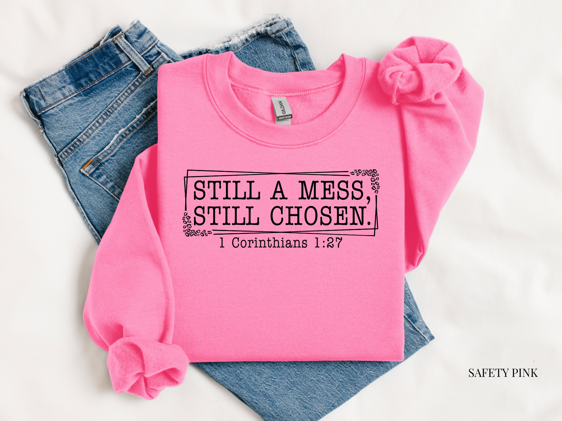 Pink sweatshirt with text 'STILL A MESS, STILL CHOSEN. 1 Corinthians 1:27' on a white background with jeans.