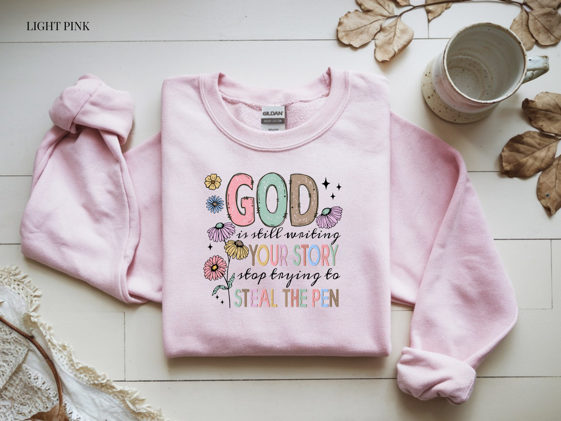 Pink sweatshirt with colorful text and flowers on a light wooden surface with a mug and leaves.