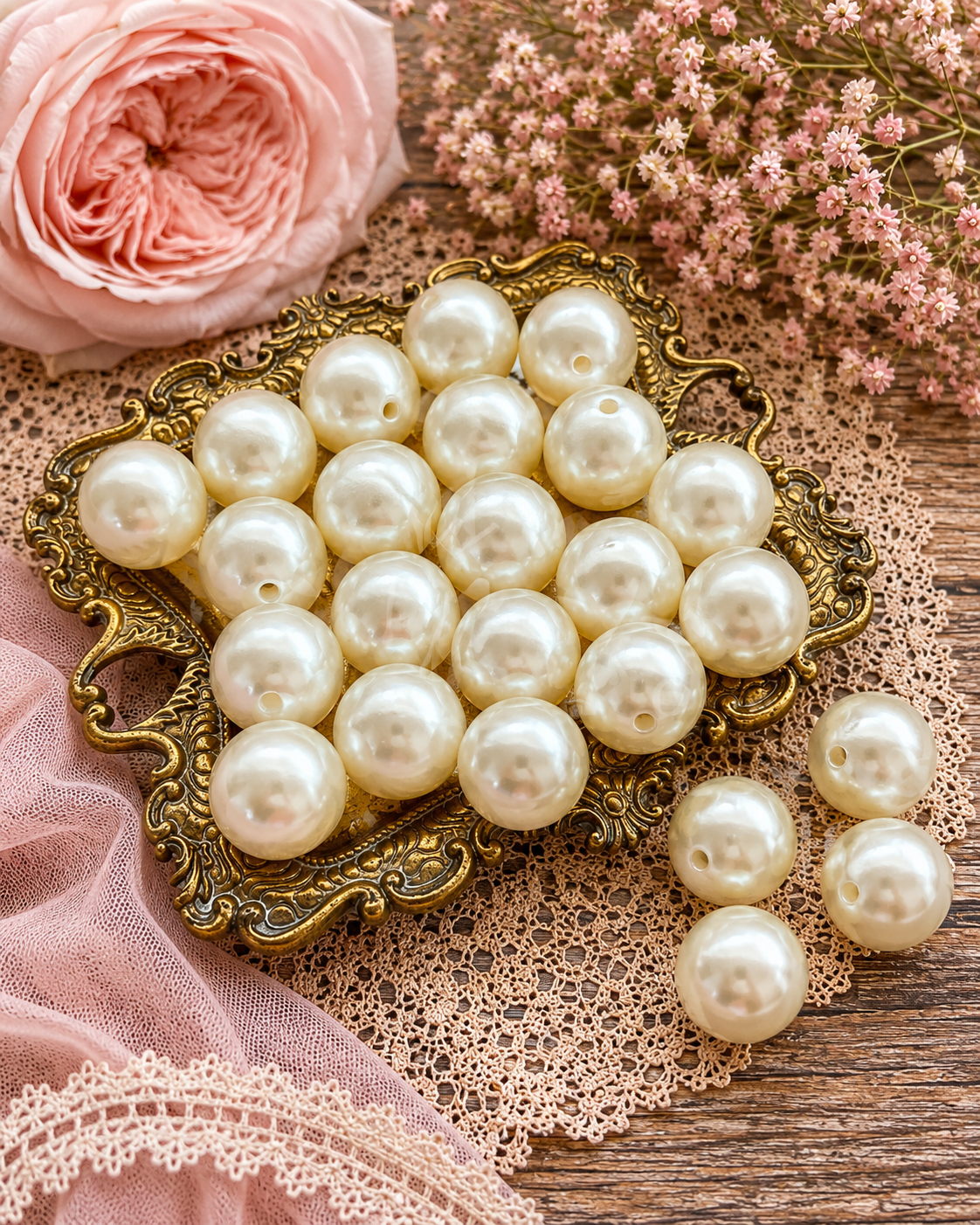 A set of round, cream-colored faux pearl beads with a single hole, displayed on an ornate gold tray with additional loose beads on a lace mat, styled with a pink rose and dried flowers.