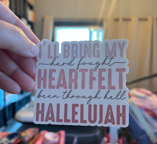 Hand holding a sticker with text 'I'll bring my hard fought heartfelt been through hell Hallelujah' in a store setting.