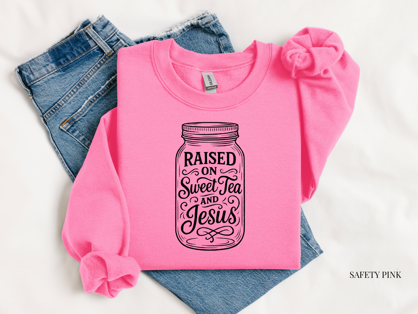 Pink sweatshirt with 'Raised on Sweet Tea and Jesus' design on blue jeans