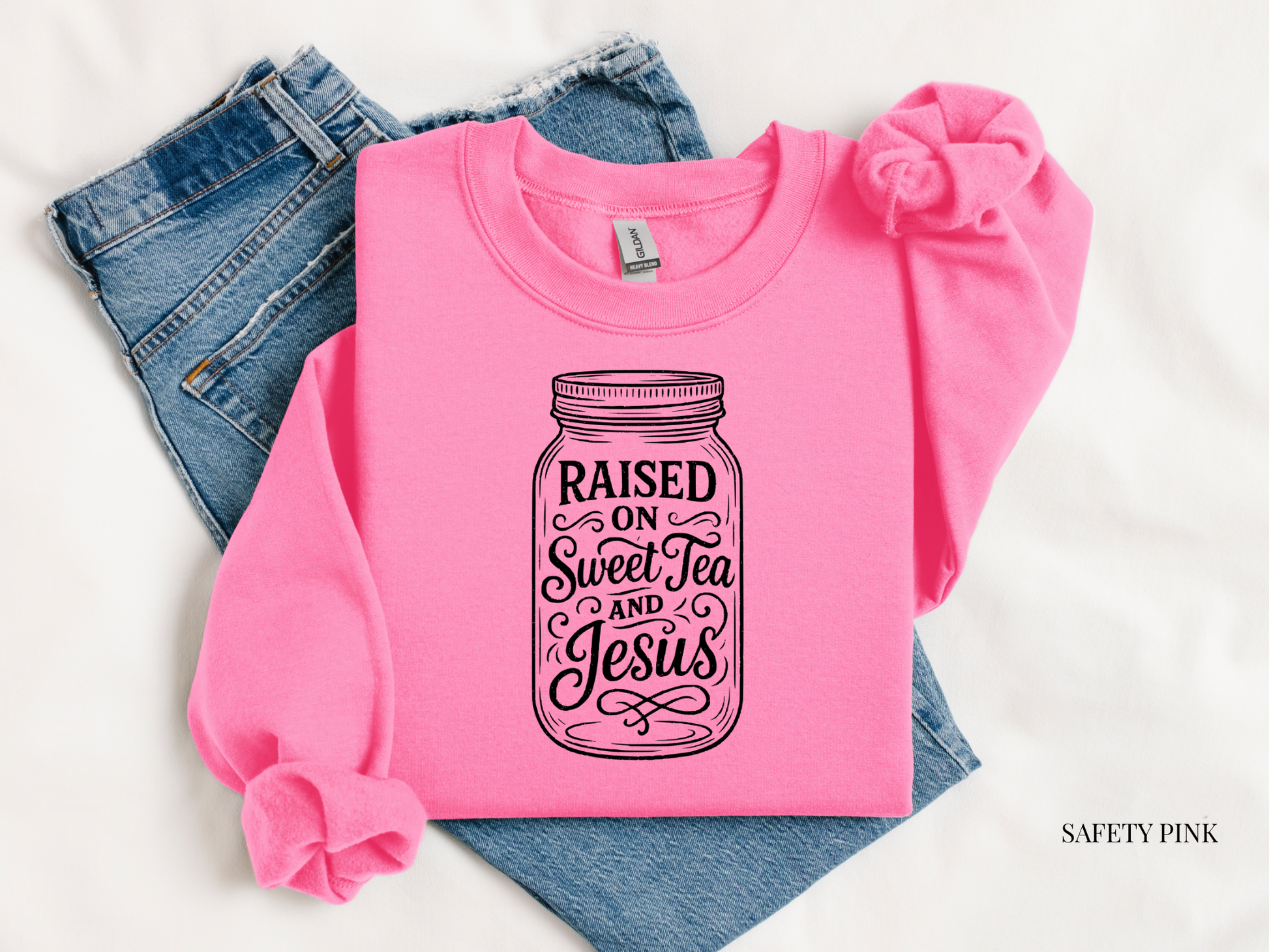 Pink sweatshirt with 'Raised on Sweet Tea and Jesus' design on blue jeans