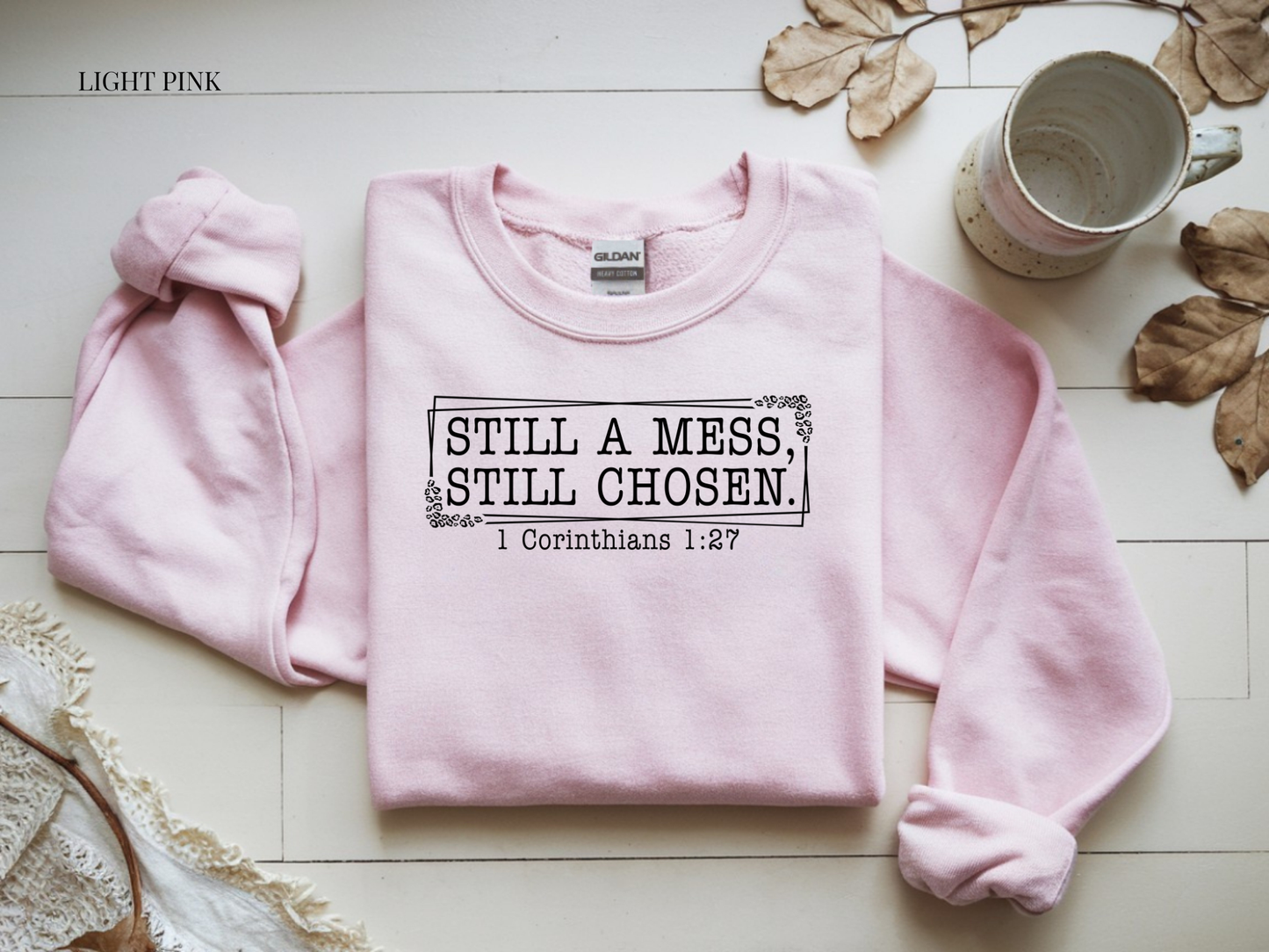 Light pink sweatshirt with text on a white surface with leaves and a mug.