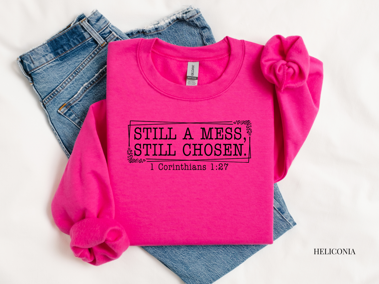 Pink sweatshirt with text 'STILL A MESS, STILL CHOSEN 1 Corinthians 1:27' on a white background