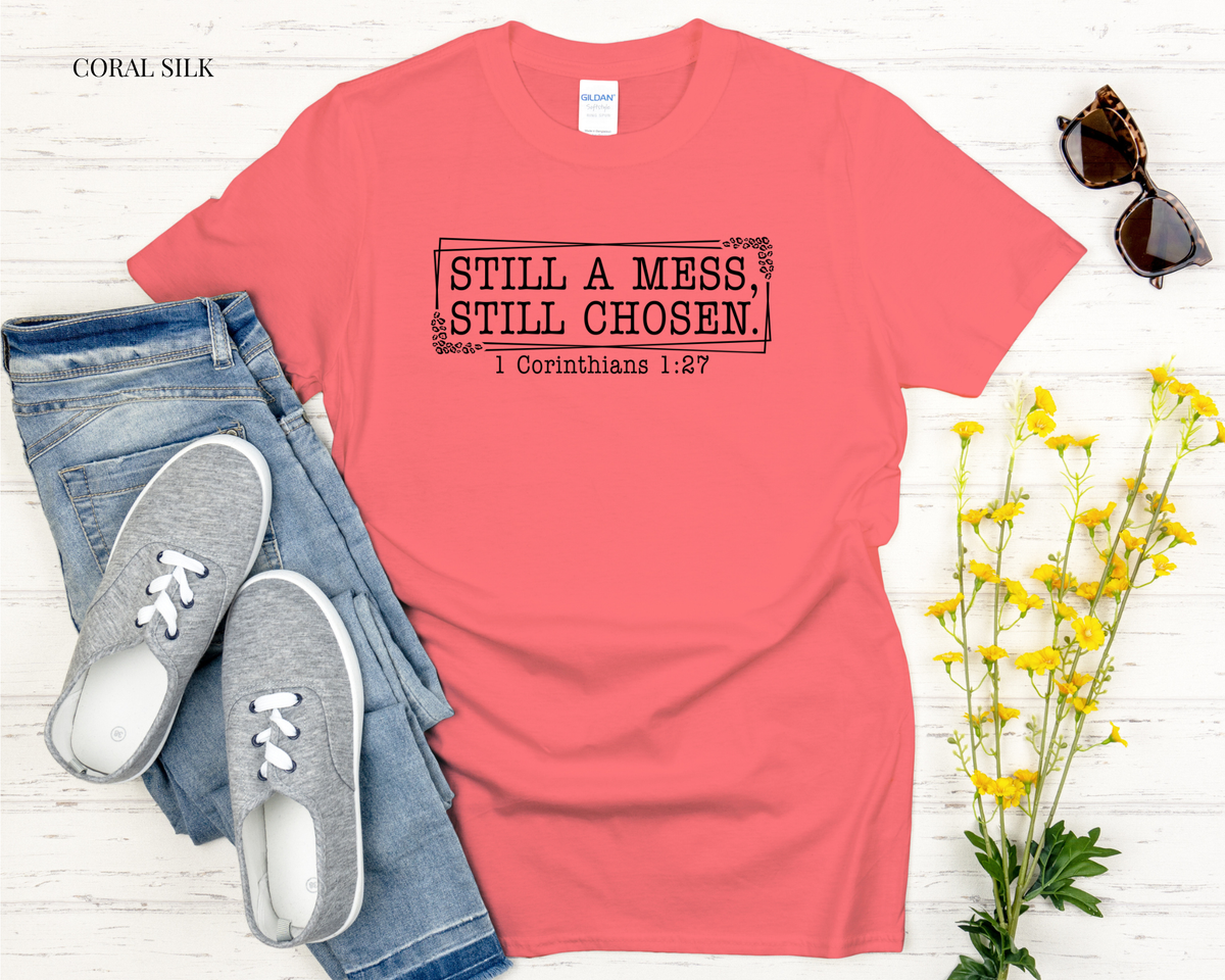 Coral t-shirt with 'STILL A MESS STILL CHOSEN' text, paired with jeans and sneakers on a light wooden surface.