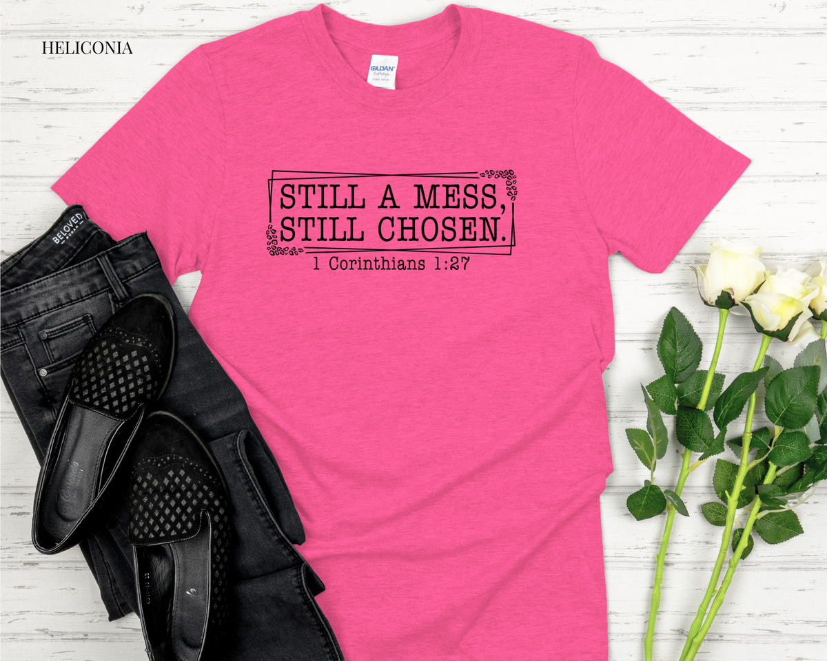 Pink t-shirt with 'Still a mess, still chosen' text on a wooden surface with black shoes and white roses.