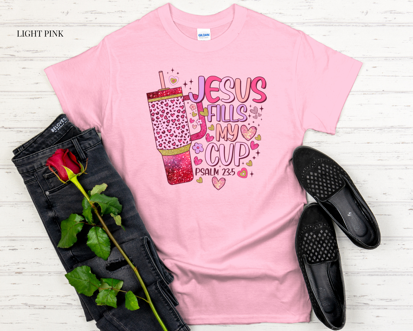 Light pink t-shirt with graphic design, black jeans, black shoes, and a rose on a light wooden surface.