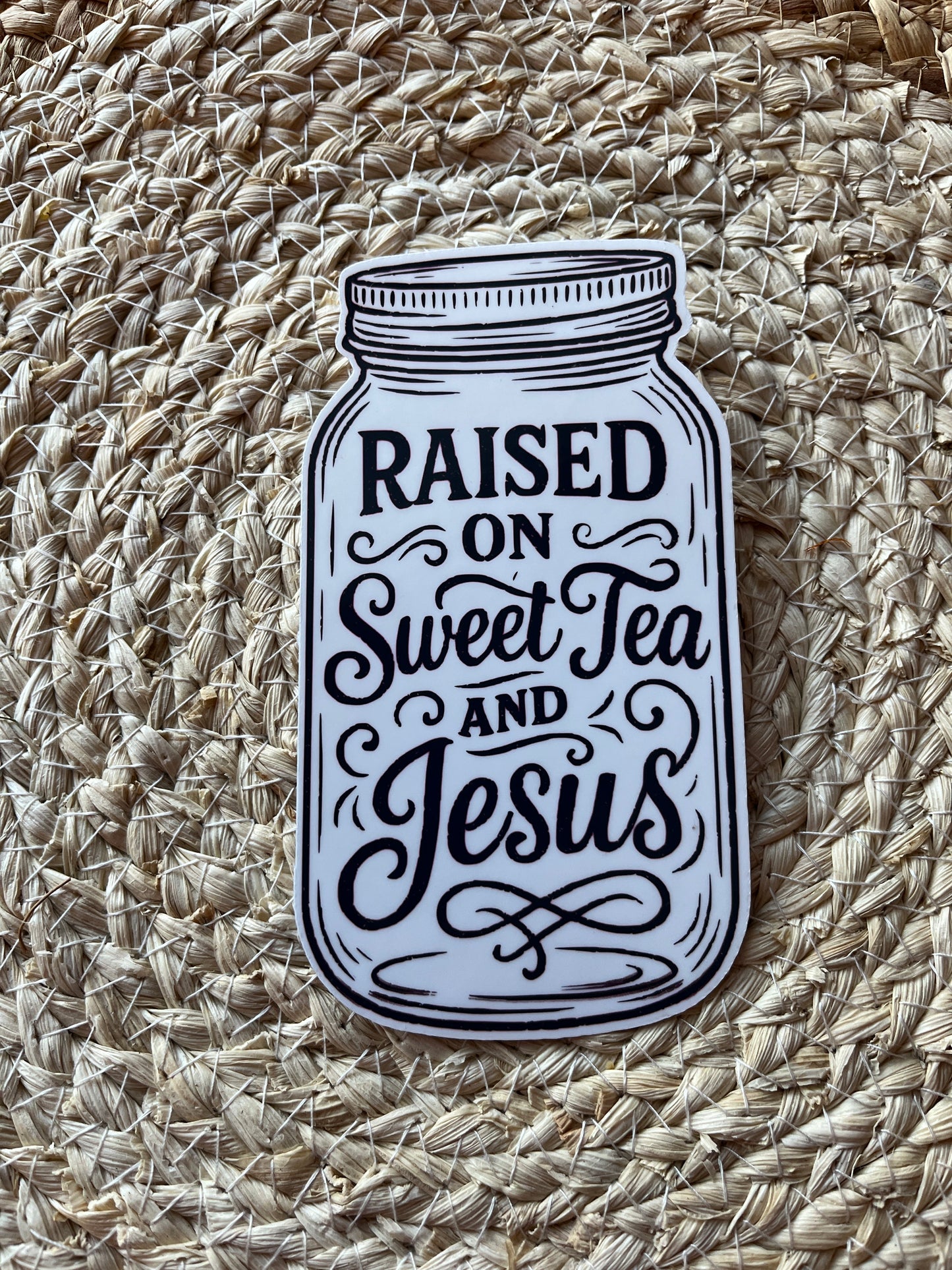 Sticker of a mason jar with text 'Raised on Sweet Tea and Jesus' on a woven mat.