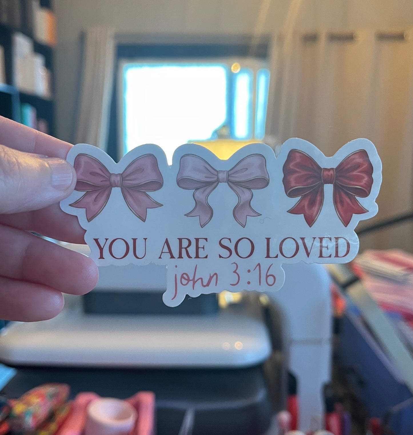 Sticker with three bows and 'You are so loved' text held by a hand in an indoor setting.