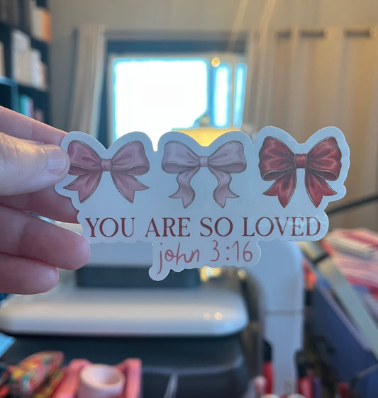 Sticker with three bows and 'You are so loved' text held by a hand in an indoor setting.