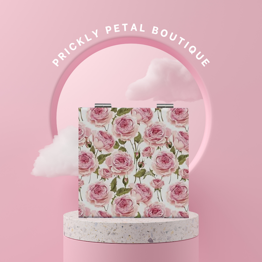 A compact mirror with a floral design, predominantly featuring pink roses, displayed on a pedestal with a branded circular backdrop.