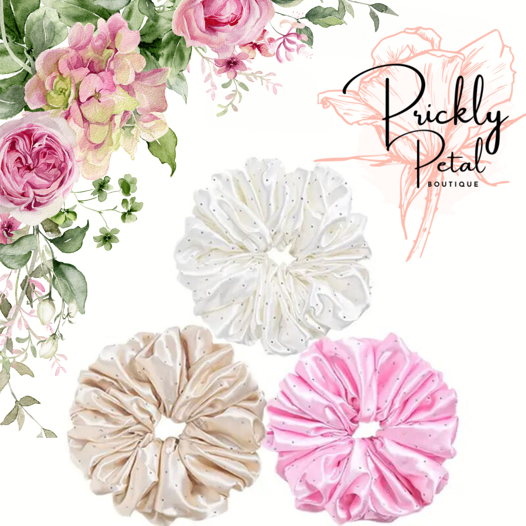 A collection of oversized rhinestone scrunchies in beige, white, and pink, displayed on a floral background.