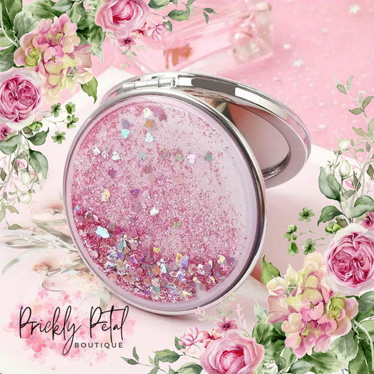 A round glitter compact mirror with a pink base and multicolored glitter, alongside heart-shaped confetti. The mirror is open, showing a dual-sided design with a magnified mirror on one side.
